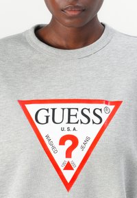 Grijze sweatshirt met een groot rood driehoeklogo en de woorden "GUESS U.S.A." en een vraagteken, wat een casual stijl en merk aangeeft.