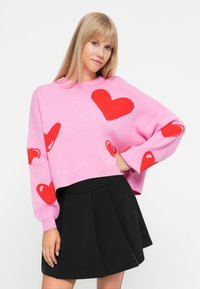 Pink sweater with red heart patterns, cropped design, ribbed cuffs, and crew neckline. Paired with a black pleated skirt.