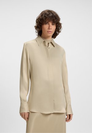 Donna con capelli ricci che indossa una camicia lunga a maniche corte beige con bottoni sopra una gonna abbinata, su uno sfondo chiaro e semplice.