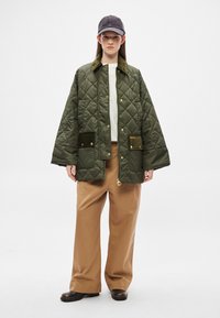 Zöld steppelt dzseki gombos záródással, két első zsebbel és puha gallérral; bézs bőszárú nadrággal és sötét sapkával párosítva.