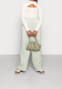 Femme portant un pantalon large vert clair, un haut à manches longues crème avec découpe, tenant un sac à main beige et portant des sandales blanches à bout ouvert.