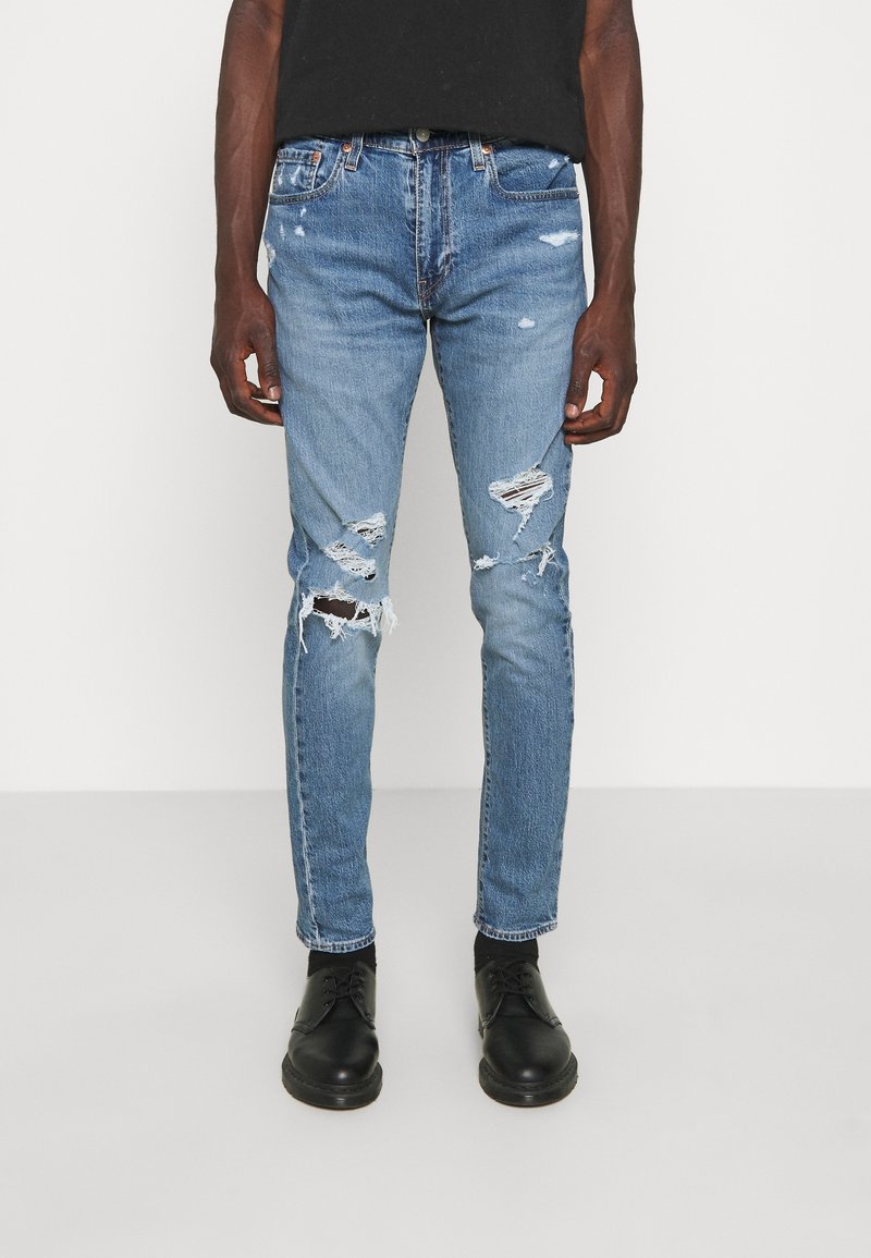 Blå denimjeans med slitna håligheter, åtsittande design och ljus tvätt. Bärs med svarta snörskor och en svart skjorta.