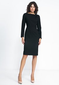 Czarna, dopasowana sukienka z długimi rękawami i dekoltem łódką, z subtelnym detalem skrętu na ramieniu. Założona z cielistymi szpilkami.