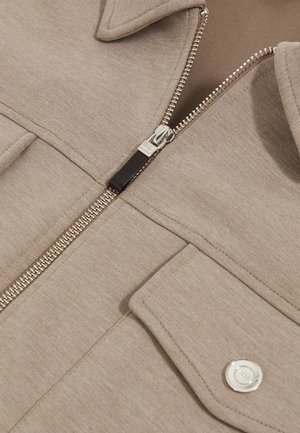 Beige Reißverschlussjacke mit glatter Textur, spitzem Kragen, silbernen Beschlägen und Klappentaschen. Der Reißverschluss verläuft zentral an der Vorderseite.