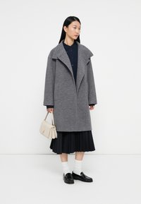 Grijze wollen jas met brede revers, oversized pasvorm en zijzakken, gedragen over een donkerblauwe blouse en een geplooide zwarte rok. Accessoires zijn een licht handtas en loafers.