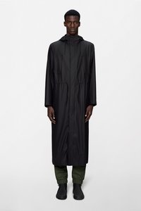 Άνδρας που στέκεται με το πρόσωπο προς τα εμπρός, φοράει μακρύ μαύρο αδιάβροχο με κουκούλα, πράσινο παντελόνι με ελαστικές μανσέτες και μαύρα παπούτσια χωρίς κορδόνια.