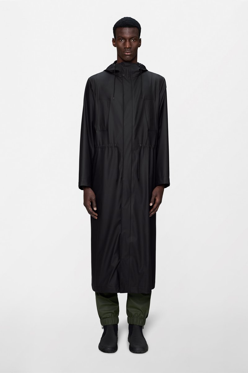 Άνδρας που στέκεται με το πρόσωπο προς τα εμπρός, φοράει μακρύ μαύρο αδιάβροχο με κουκούλα, πράσινο παντελόνι με ελαστικές μανσέτες και μαύρα παπούτσια χωρίς κορδόνια.