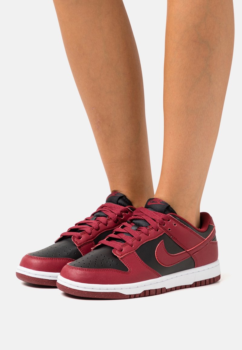 Baskets basses Nike rouges et noires avec semelles blanches portées sur des jambes nues sur fond blanc.