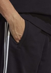 Juodos sportinės šortai su baltais šonų raiščiais, priekinėje kišenėje matoma ranka. Medžiaga atrodo minkšta ir rūdinga.