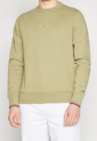 Felpa verde oliva con scollatura a girocollo, caratterizzata da un piccolo logo bianco "Tommy Hilfiger" sul petto e dettagli a righe sui polsini.