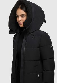 Čierna puffová bunda s hrubou kapucňou s fleecovou podšívkou. Obsahuje zips na prednej strane, bočné vrecká a nášivku s logom na rukáve.
