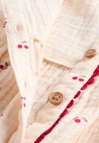 Chemise en tissu crème clair ornée de broderies de cerises rouges, boutons en bois, et un accent froncé bordeaux le long de la patte de boutonnage.