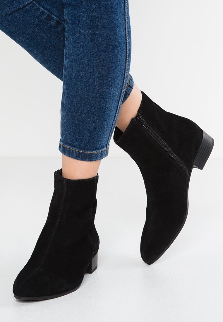 Bottines en daim noir avec fermeture éclair latérale, portées avec un jean denim bleu coupé au-dessus de la cheville, sur fond clair.
