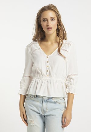Jonge vrouw met lang haar draagt een witte blouse met knopen en kanten details en lichte distressed jeans tegen een effen lichte achtergrond.