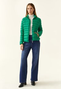 Zelena puffer jakna s horizontalnim šavovima, visokim ovratnikom i zatvaranjem na patentni zatvarač. Nosena preko bijelog turtleneck-a i uparena s plavim širokim trapericama.