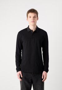 Μαύρο μακρυμάνικο πόλο μπλουζάκι με κολάρο, τρεις κουμπάκια και διακριτό λογότυπο στο αριστερό στήθος, συνδυασμένο με σκούρα τζιν.