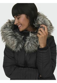 Veste en duvet noire avec un grand col en fausse fourrure grise, fermeture zippée, poignets côtelés et motif matelassé sur les manches.