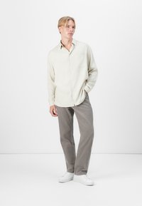 Helles beiges Hemd mit Knopfleiste und einer Brusttasche, kombiniert mit grauen Cordhosen und weißen Sneakers; das Modell steht vor einem neutralen Hintergrund.