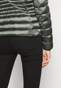 Grön puffyjacka med quiltat mönster, med en rund logotypapplikation. Svarta jeans synliga nedanför, med enkla bakfickor.