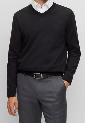 Mężczyzna ubrany w czarny sweter z dekoltem w serek na białą koszulę, szare spodnie z czarnym paskiem, ręka w kieszeni na jednolitym tle.