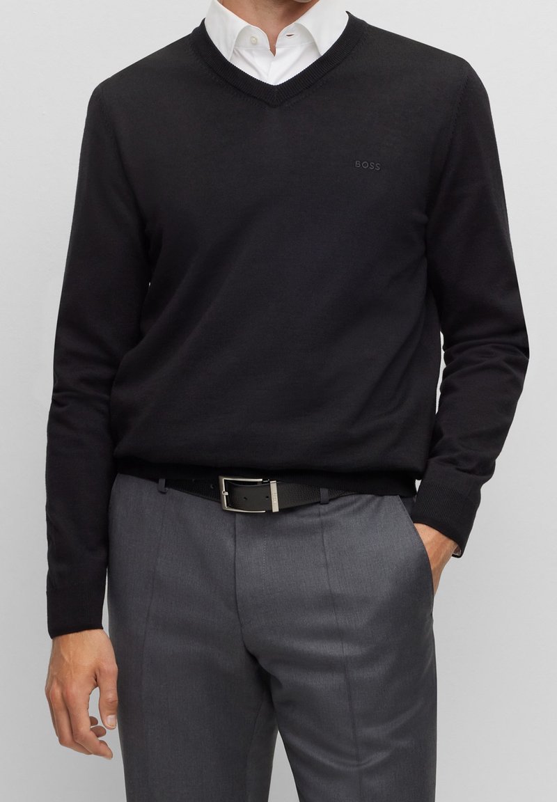Homme portant un pull noir à col en V par-dessus une chemise blanche, un pantalon gris avec ceinture noire, main dans la poche sur un fond uni.