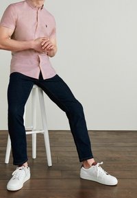 Korte mouwen, lichtroze overhemd met knopen en een klein logo, gecombineerd met donkere jeans en witte sneakers, zittend op een witte kruk.