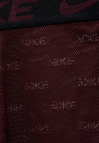 Σκούρο μπορντό πλεκτό ύφασμα με επαναλαμβανόμενες μικρές τρύπες και αχνά ανάγλυφα λογότυπα "NIKE" σε διακριτό μοτίβο.