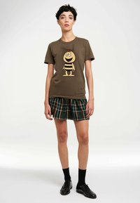 Braunes Baumwoll-T-Shirt mit einem Cartoon-Bienen-Design. Kombiniert mit einem grünen, karrierten Faltenrock. Schwarze Schuhe und Socken vervollständigen das Outfit.
