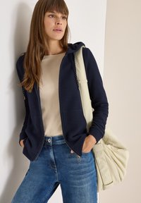 Donna che indossa una felpa blu navy con zip, un top beige, jeans blu e porta una grande borsa trapuntata color crema sulla spalla, in piedi contro un muro.