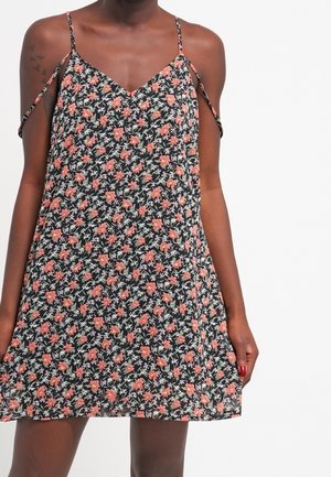 Frau trägt ein schwarzes, ärmelloses Blumenkleid mit dünnen Trägern und schulterfreien Details und steht vor einem schlichten weißen Hintergrund.