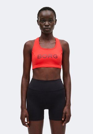 Athletische Frau trägt einen roten Sport-BH mit dem Text "BORG" und schwarze, hoch taillierte Bikeshorts, steht vor einem schlichten hellen Hintergrund.