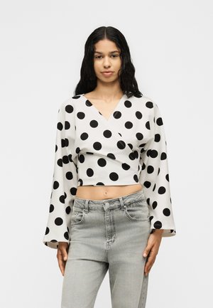 Γυναίκα με μακριά σκούρα μαλλιά, φοράει λευκό cropped πουκάμισο τύπου wrap με μαύρες πουά και ανοιχτό γκρι τζιν με ψηλή μέση, στέκεται μπροστά σε λευκό φόντο.