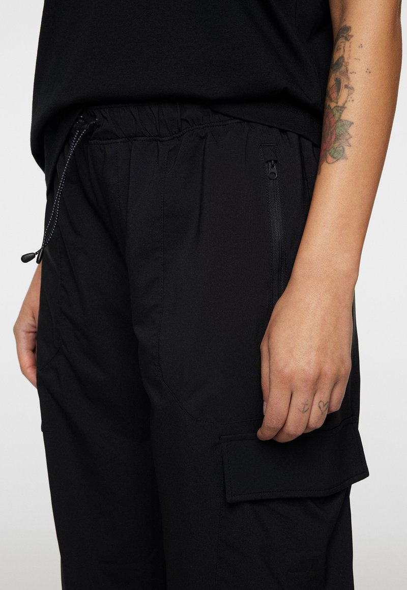 Pantaloni cargo neri con vita regolabile tramite cordoncino, tasca laterale con zip e tasca a toppa sulla coscia. Tessuto dalla texture liscia.