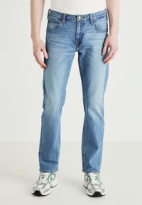 Ljusblå denimjeans med rak passform, fem fickor och subtilt slitage. Bärs med grå sneakers med gröna detaljer.