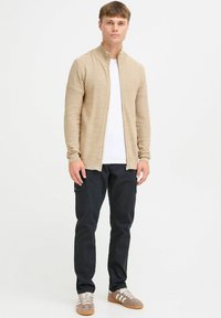 Chaqueta de punto beige con cremallera sobre una camisa blanca, combinada con pantalones cargo oscuros y zapatillas grises. Diseño minimalista con detalles prácticos.