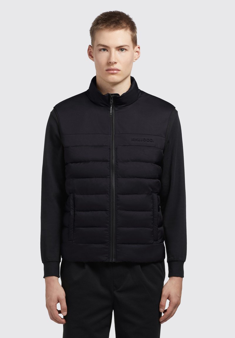 Sort ærmeløs puffer vest med lynlås foran, der har en quiltet tekstur og en høj krave. Glat stof på skuldrene og ryggen.