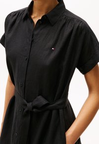Rochie neagră din material texturat, cu închidere cu nasturi în față, mâneci scurte și o curea legată la talie. Accent logo mic pe piept.