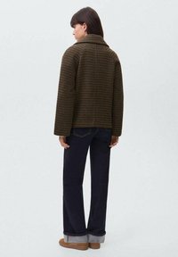 Cappotto a quadri marrone e nero con design doppiopetto, colletto e maniche larghe, abbinato a jeans blu scuro e scarpe beige.
