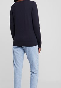 Kvinna iklädd en enfärgad marinblå långärmad tröja och ljusblå högmidjade jeans, stående med ryggen mot kameran.