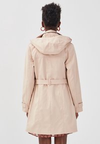 Vrouw met krullend haar, gekleed in grote hoepeloorbellen en een beige trenchcoat met capuchon en riem, staat met haar rug naar de camera op een witte achtergrond.