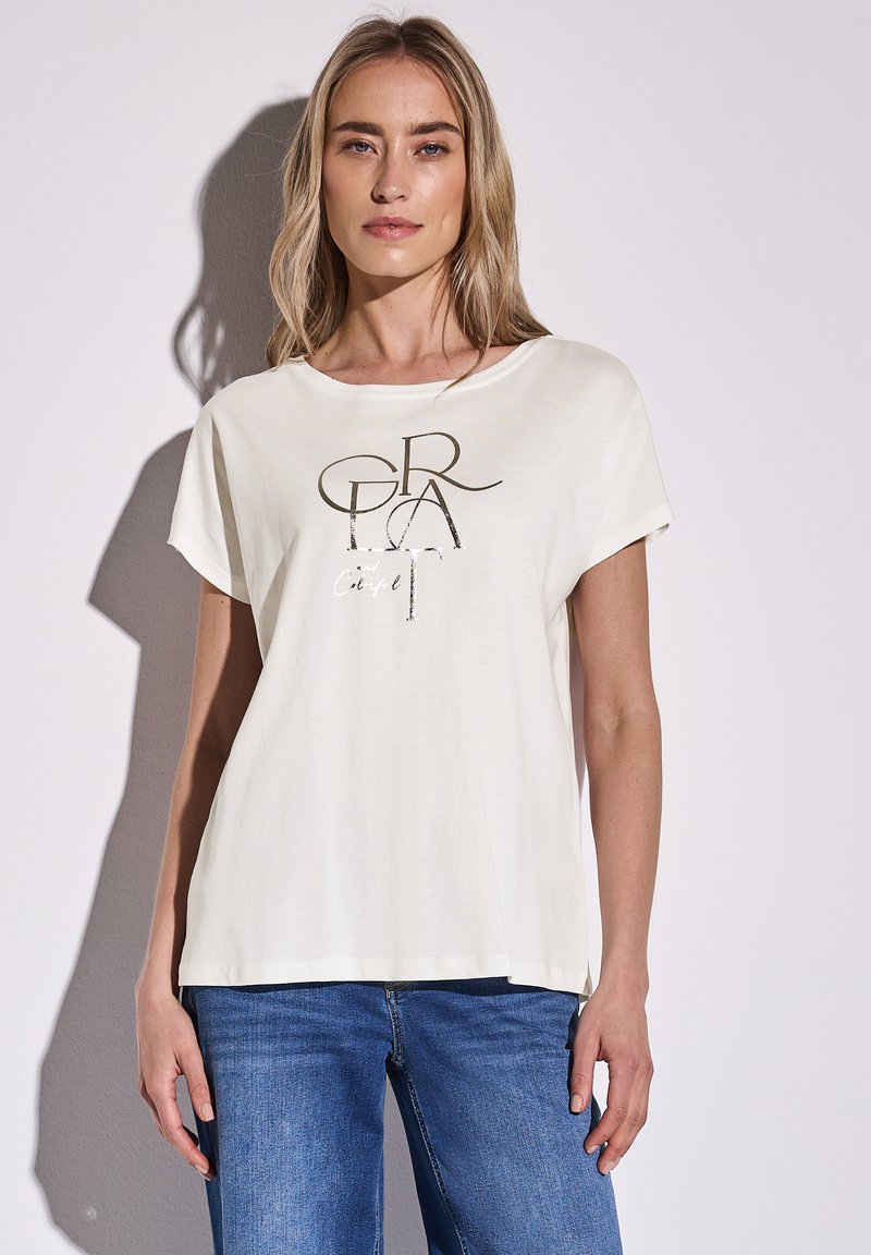 Lichtcrème t-shirt met een losse pasvorm, korte mouwen, ronde halslijn, met een metalen grafische print op de voorkant. Draagt met blauwe jeans.