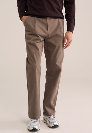 Mann, der braune gerade geschnittene Hose, bordeauxfarbenes Langarmoberteil und weiße Sportschuhe trägt, steht mit einer Hand in der Tasche.