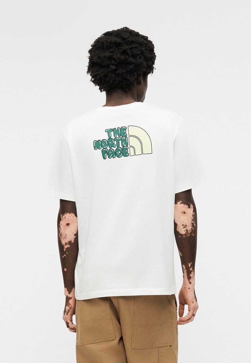 Άτομο με σγουρά μαλλιά και λεύκη που φοράει λευκό μπλουζάκι με πράσινο λογότυπο "The North Face" και καφέ παντελόνι, φωτογραφημένο από πίσω.