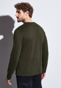 Camisa de manga larga verde oscuro con cuello redondo, textura suave y detalles de costura visibles. Representada desde la parte posterior, mostrando el ajuste.