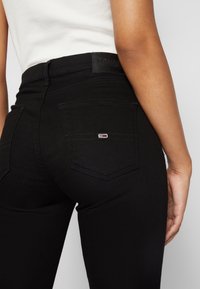 Persona que lleva puesto unos vaqueros negros ajustados con un logo blanco, rojo y azul en el bolsillo trasero y una camiseta sin mangas blanca.