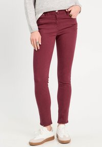 Jean skinny bordeaux en mélange de coton, présentant une texture lisse, une taille mi-haute et des poches devant. Portés avec des baskets blanches.
