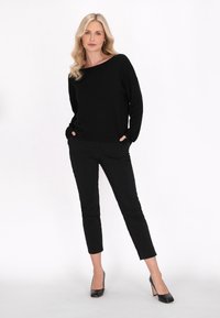 Fekete kötött pulóver, széles nyakkivágással, fekete slim-fit nadrággal és fekete magassarkú cipővel párosítva, fehér háttér előtt.