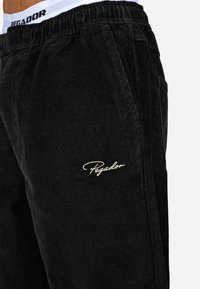 Czarne spodnie ze sztruksu z teksturowaną powierzchnią, elastycznym pasem w białe i czarne logo oraz dyskretnym złotym haftem na lewym udzie.