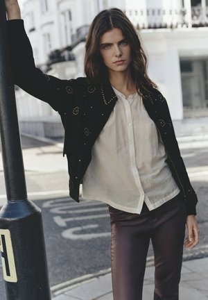 Jeune femme aux longs cheveux foncés vêtue d'une blouse blanche, d'une veste noire cloutée et d'un pantalon sombre, appuyée contre un poteau métallique noir dans une rue de la ville.