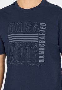 T-paita tummansinisestä puuvillasta, jossa on vaaleanharmaita printtigrfikoita, mukaan lukien teksti ja vaaka viivat. Rajoitettu erä.